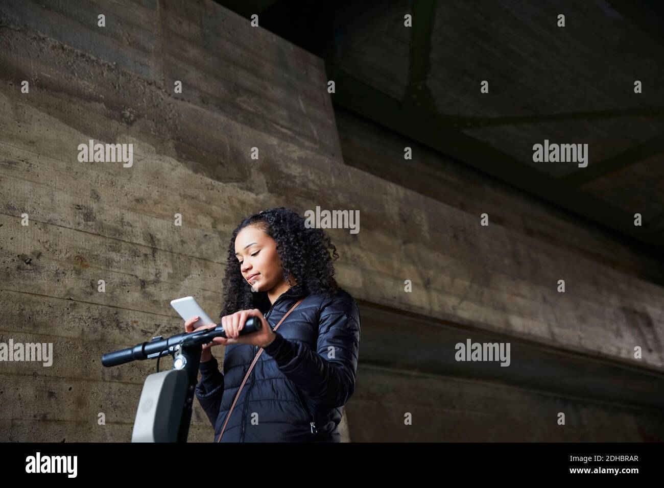 Low-Angle-Ansicht von Teenager-Mädchen mit Elektro-Push-Roller Mobiltelefon an der Wand unter der Brücke verwenden Stockfoto