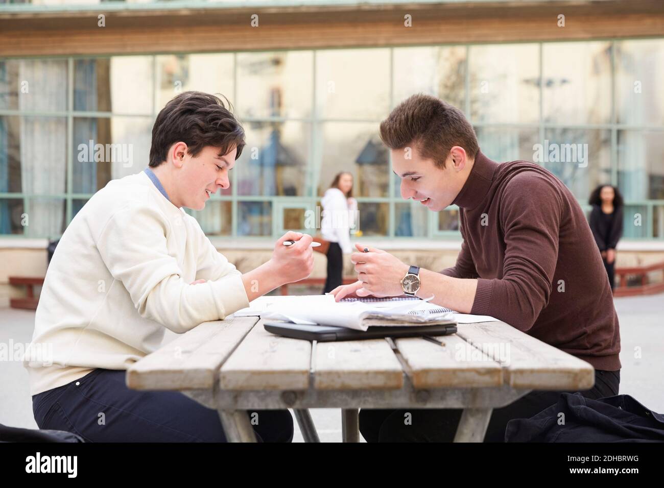 Männliche Teenager-Freunde lächeln, während sie am Tisch im Schulhof studieren Stockfoto