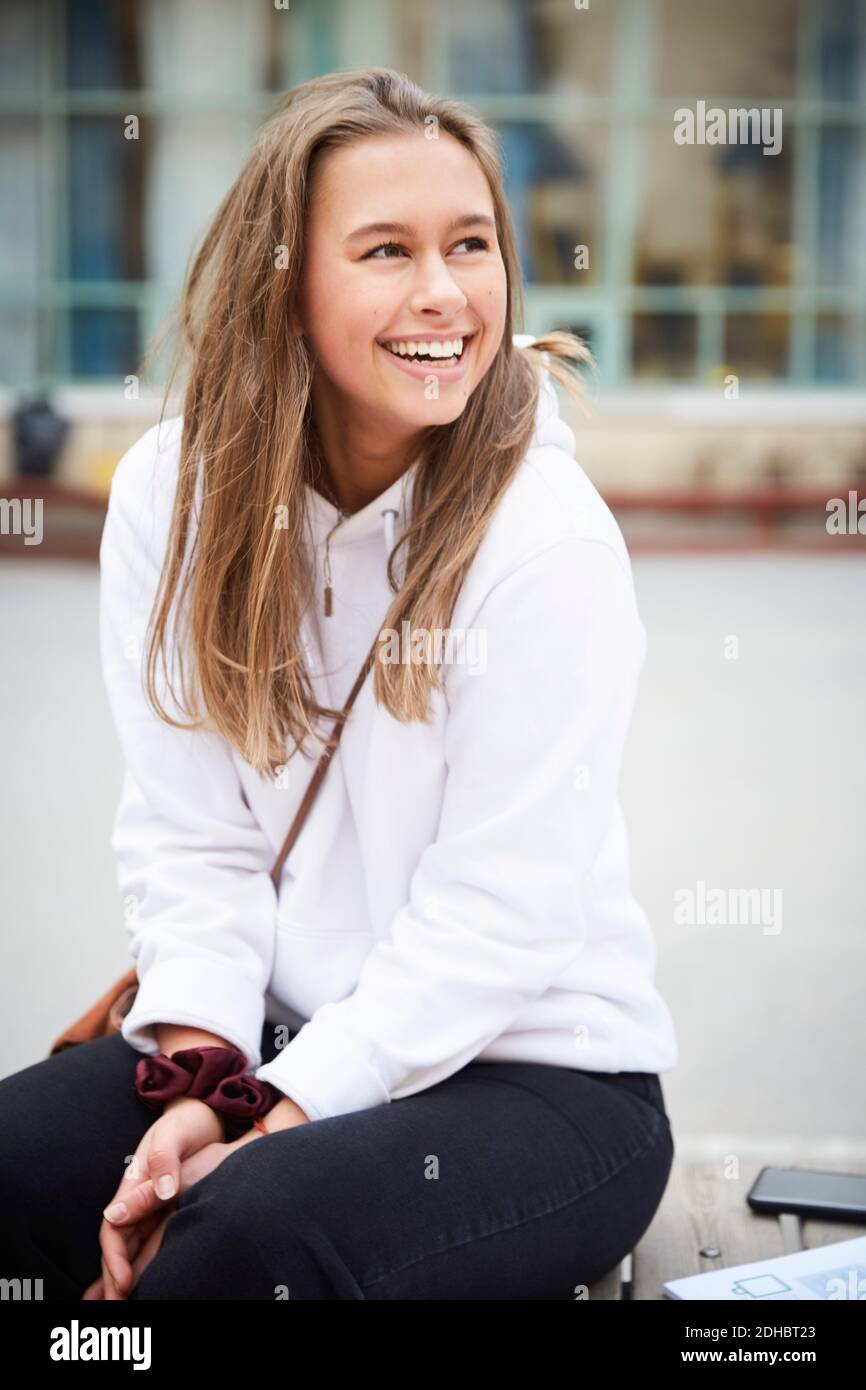 Lächelndes Teenager-Mädchen, das wegschaut, während es im Schulhof sitzt Stockfoto