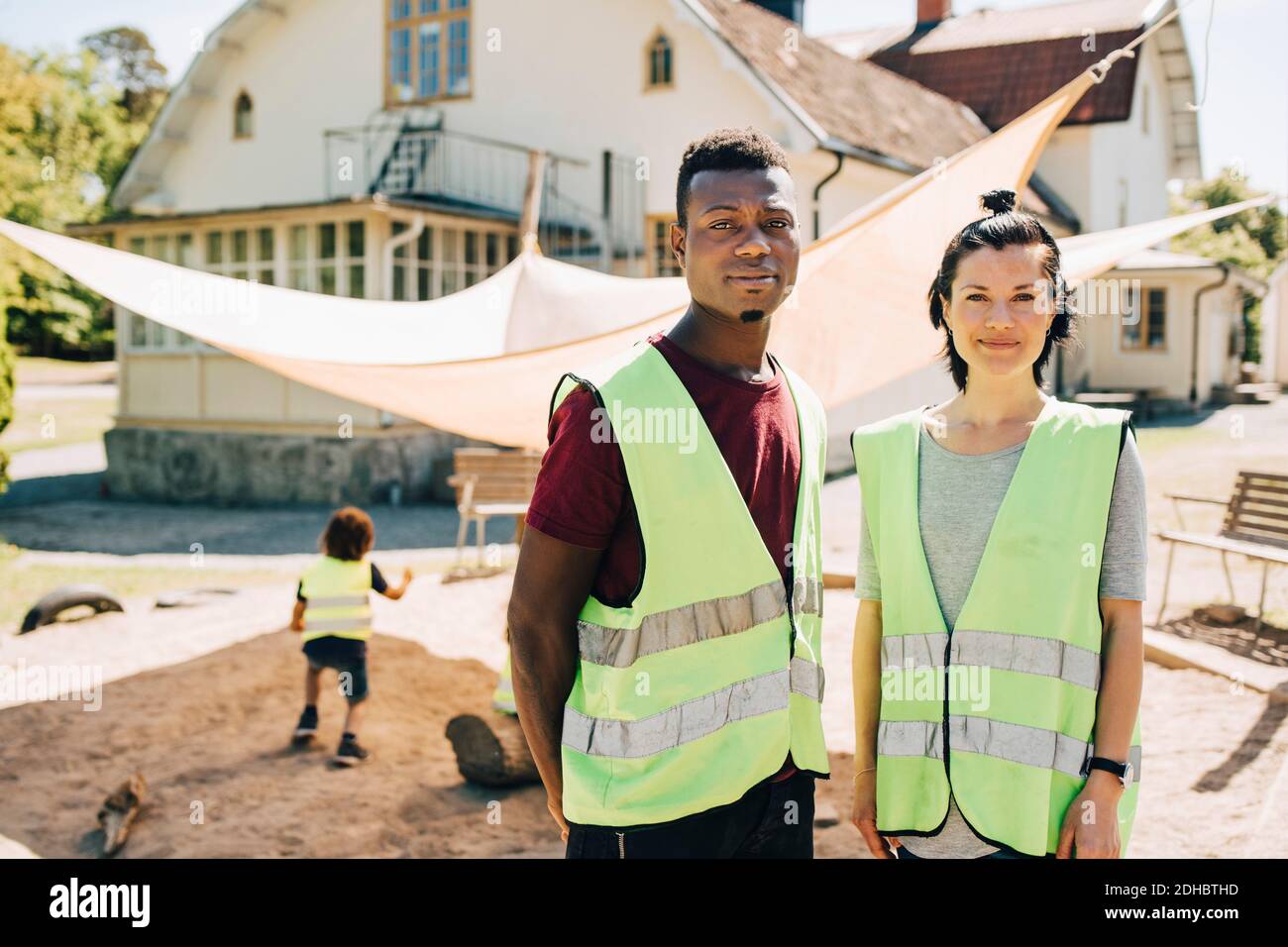 Portrait von selbstbewussten mittleren Erwachsenen männlichen und weiblichen Lehrern stehen In reflektierender Kleidung auf dem Spielplatz Stockfoto