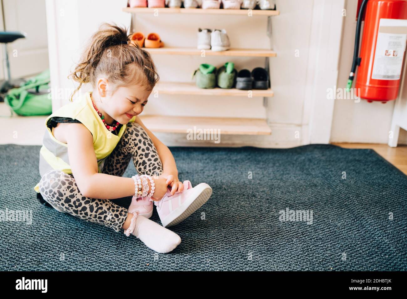 Volle Länge des Mädchens macht Gesicht, während auf dem Teppich sitzen Schuhe in der Garderobe bei der Kinderbetreuung tragen Stockfoto