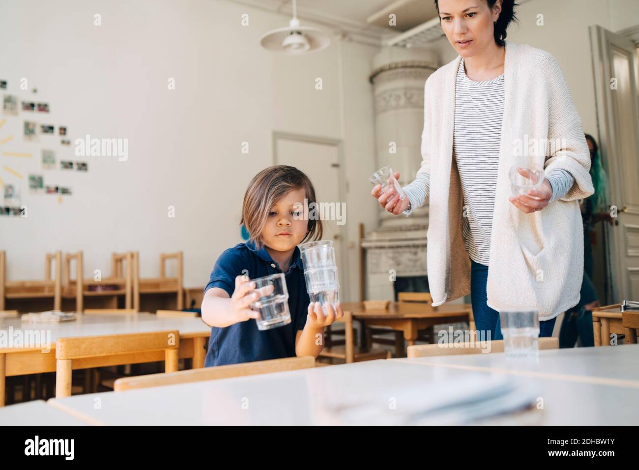 Lehrer für mittleren Erwachsenen mit Jungen, der Trinkgläser auf dem Tisch arrangiert Im Klassenzimmer bei der Kinderbetreuung Stockfoto