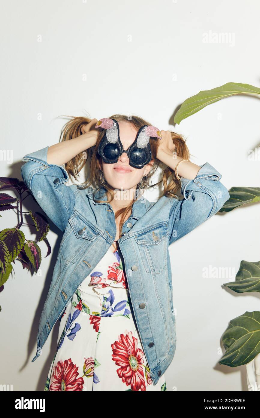 Lächelnde junge Frau mit Händen im Haar, die inmitten von Pflanzen stehen Gegen weiße Wand zu Hause Stockfoto