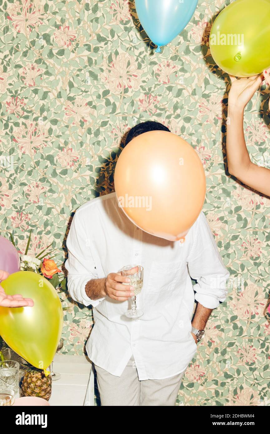 Verschwomenes Gesicht eines jungen Mannes, der beim Stehen einen Drink hält Frau mit Luftballons gegen Tapete zu Hause während des Abendessens p Stockfoto