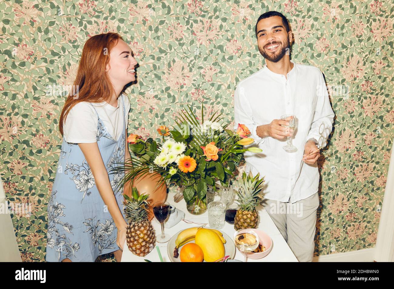 Lächelnde junge Freunde, die beim Abendessen am Tisch gegen die Tapete stehen Party zu Hause Stockfoto