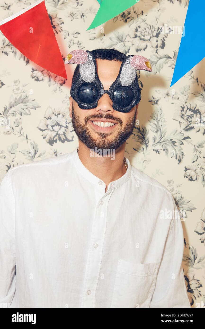 Lächelnder junger Mann, der eine Brille trägt, während er gegen die Tapete steht Wohnung während der Party Stockfoto