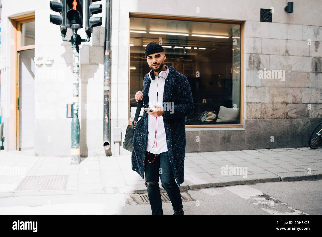 Porträt des selbstbewussten jungen Geschäftsmann zu Fuß mit Smartphone und Tasche gegen Gebäude in der Stadt Stockfoto