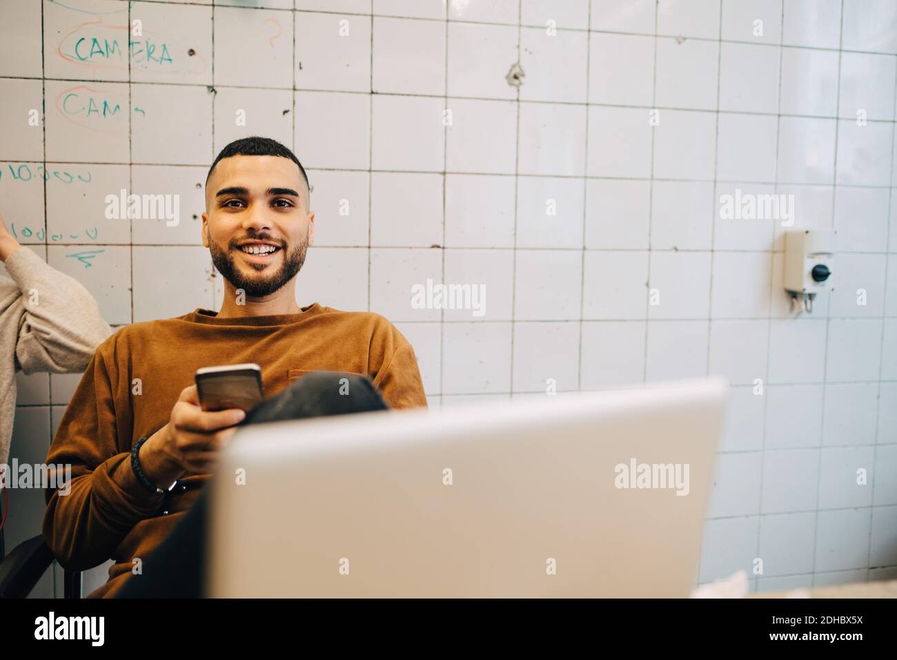 Porträt eines lächelnden jungen Geschäftsmannes, der mit einem Smartphone gegen den Tisch sitzt fliesenwand im Kreativbüro Stockfoto