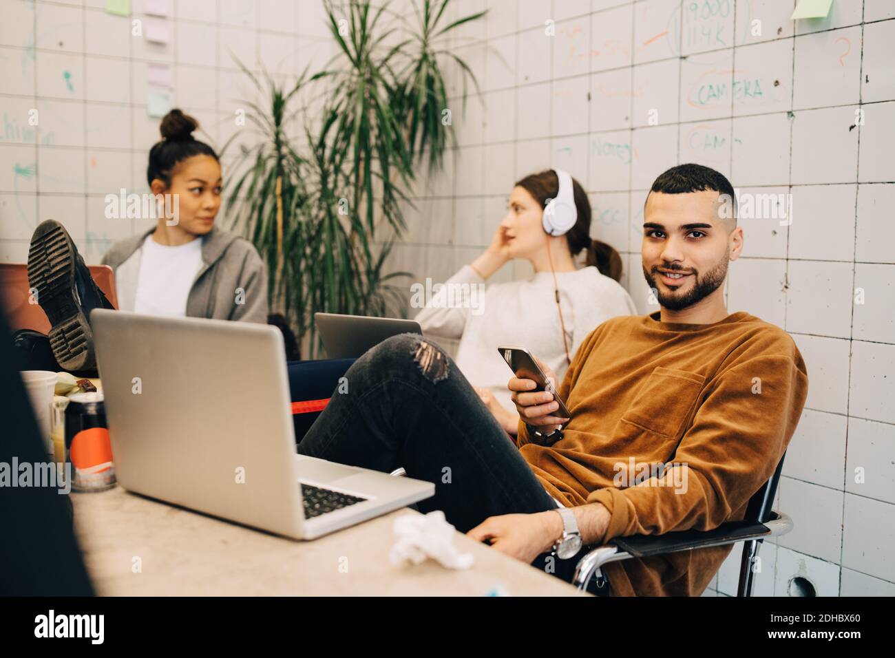 Porträt eines lächelnden jungen Geschäftsmannes, der während des Sitzens Smartphone hält Mit Kolleginnen im kleinen Büro Stockfoto