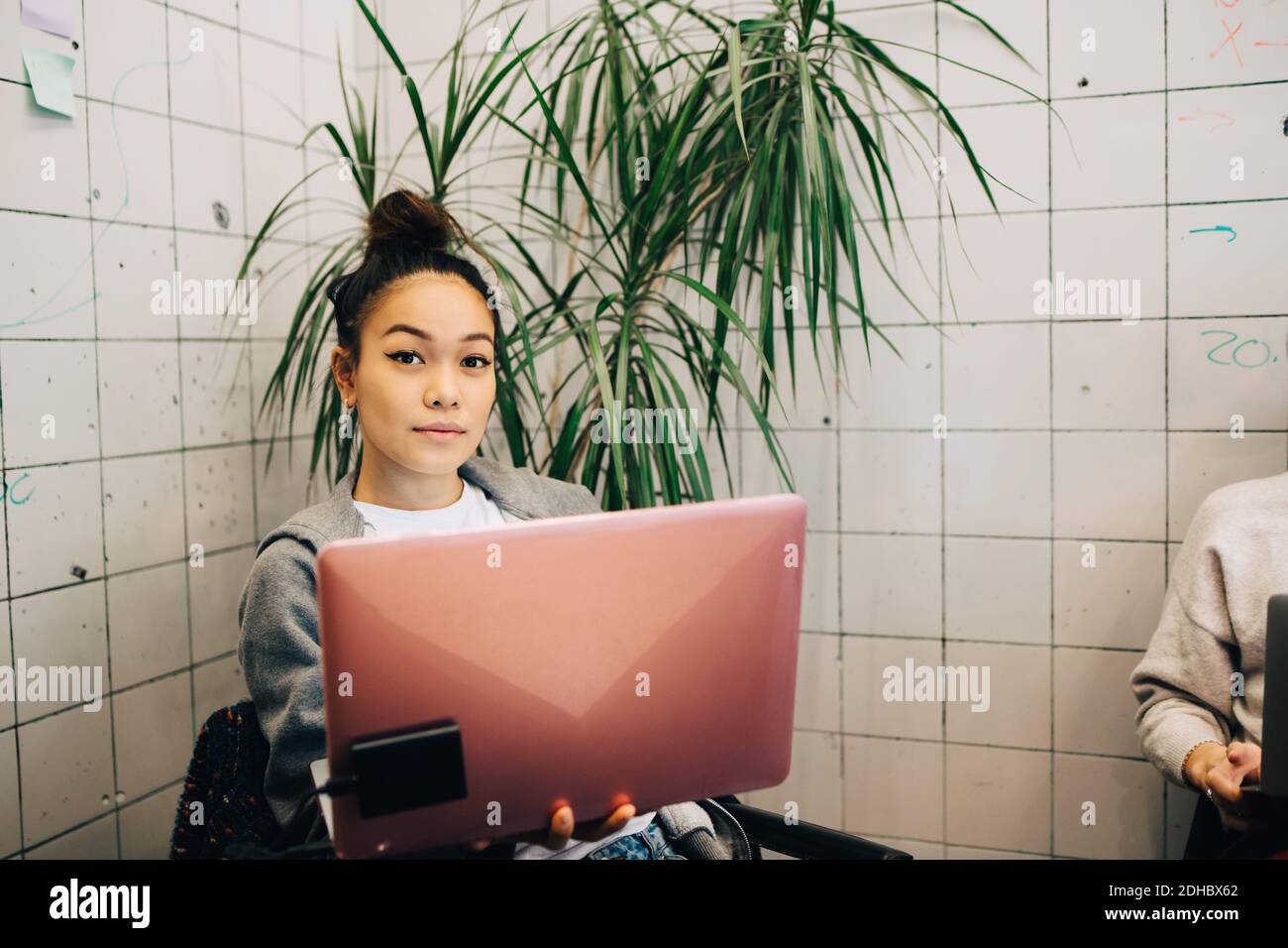 Porträt einer selbstbewussten jungen Hacker-Frau mit Laptop während des Sitzens Gegen Fliesenwand in kleinem Kreativbüro Stockfoto