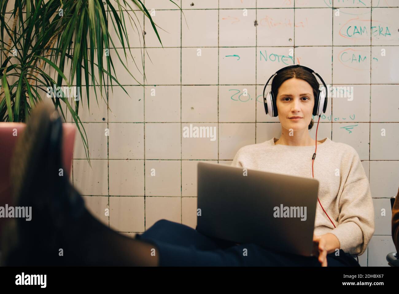Porträt einer selbstbewussten jungen Computerhackerin, die über Kopfhörer zuhört Beim Sitzen mit Laptop gegen Fliese w Stockfoto