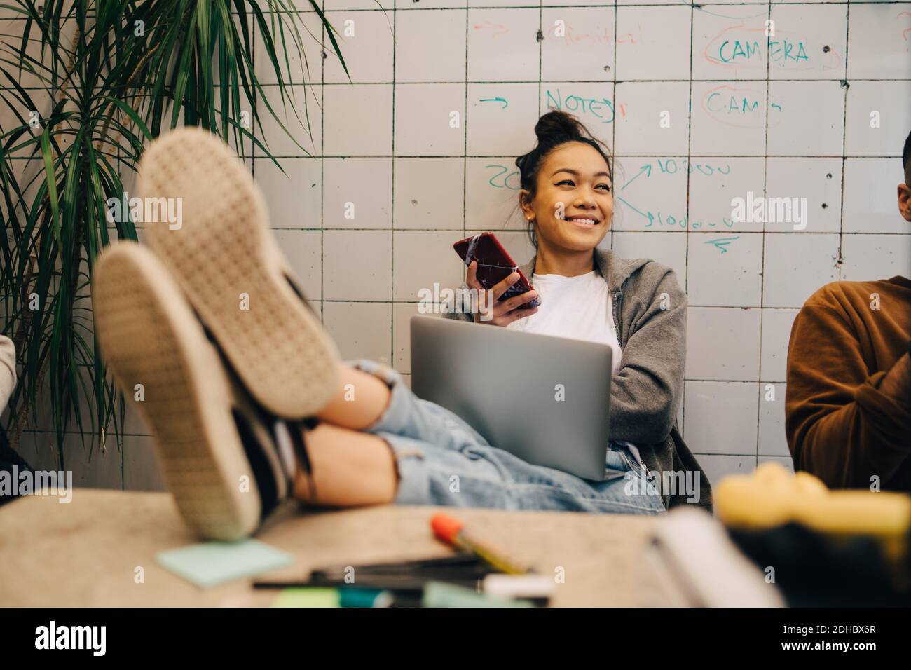 Lächelnde junge Geschäftsfrau, die mit den Füßen auf dem Schreibtisch sitzt Wireless-Technologien von Kollegen gegen die Wand im Büro Stockfoto