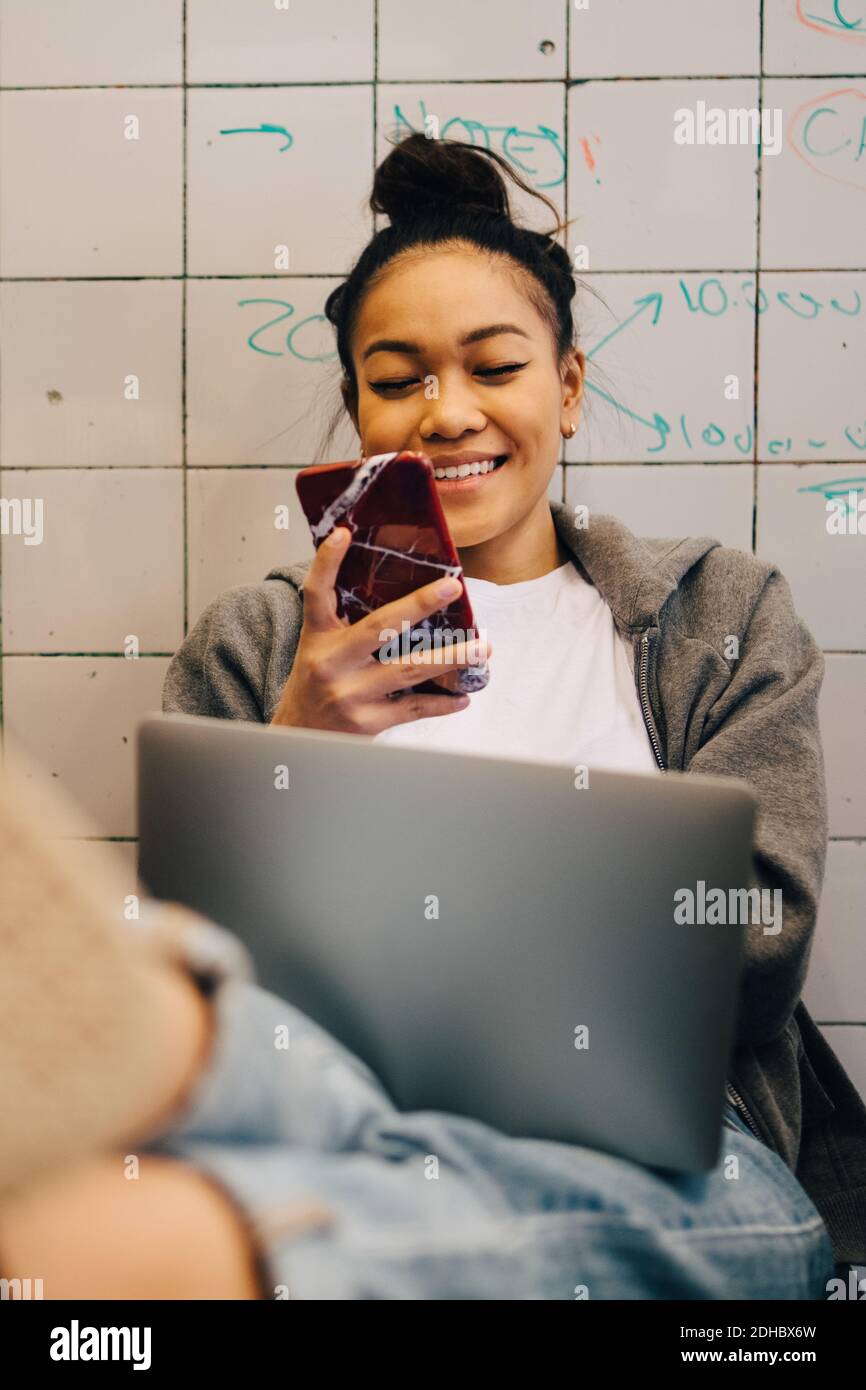 Lächelnd junge Hacker mit Smartphone, während sie mit sitzen Laptop gegen Fliesenwand im kleinen Kreativbüro Stockfoto