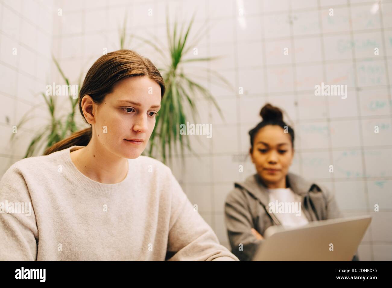 Selbstbewusste junge Geschäftsfrau mit Laptop, während sie von einer Kollegin sitzt Im kleinen Kreativbüro Stockfoto