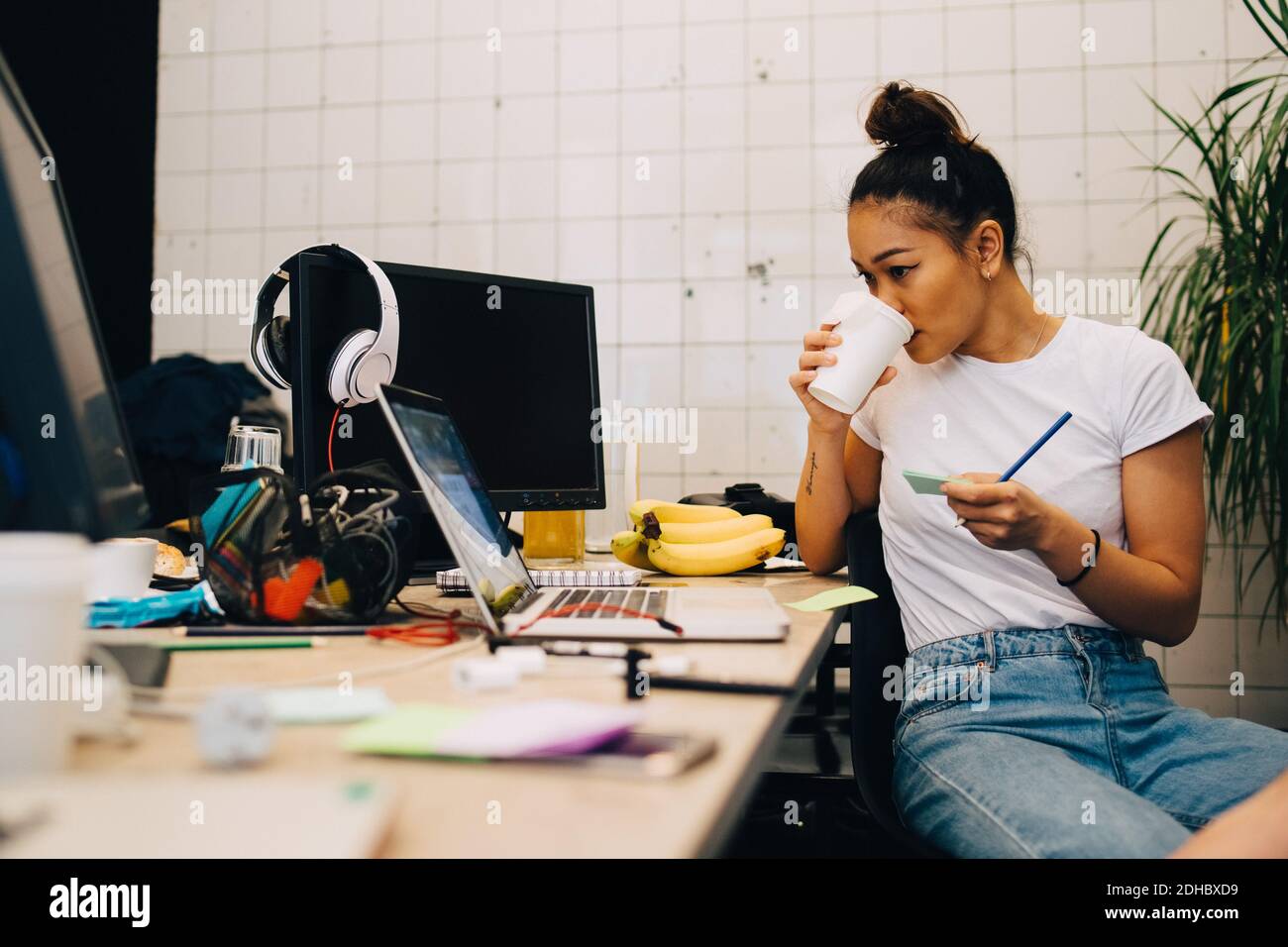 Junge Geschäftsfrau trinkt Kaffee, während sie am Schreibtisch in klein sitzt Kreatives Büro Stockfoto