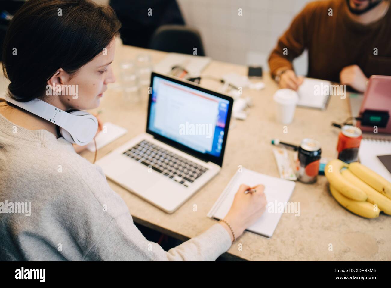 High-Angle-Ansicht der weiblichen Programmierer Schreiben auf Buch durch Laptop, während ein männlicher Kollege im kreativen Büro am Schreibtisch arbeitet Stockfoto