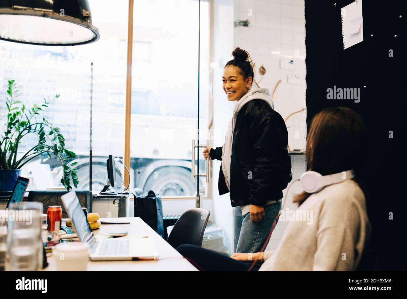 Lächelnde junge Geschäftsfrau, die die Tür hält, während sie bei einer Kollegin steht Im Kreativbüro Stockfoto