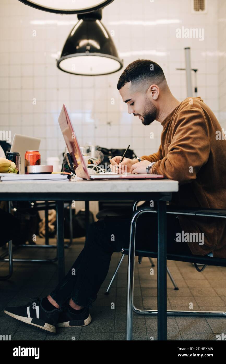 Volle Länge Seitenansicht des selbstbewussten jungen männlichen Computerprogrammierers Schreiben, während Sie mit einem Laptop am Schreibtisch sitzen, in kleiner Größe Stockfoto