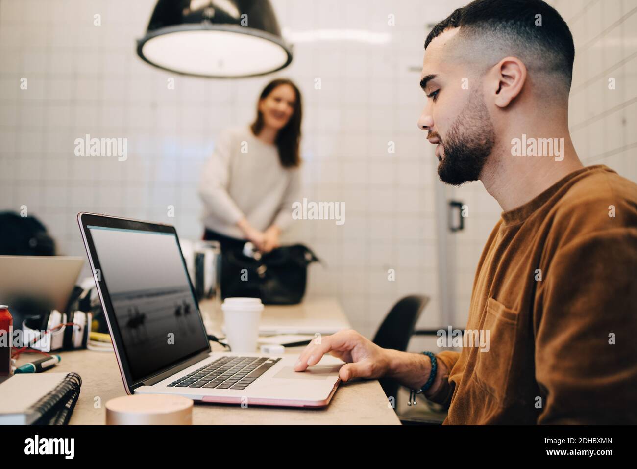 Seitenansicht des jungen männlichen Hackers mit Laptop, während weiblich Kollege, der in einem kleinen Büro arbeitet Stockfoto