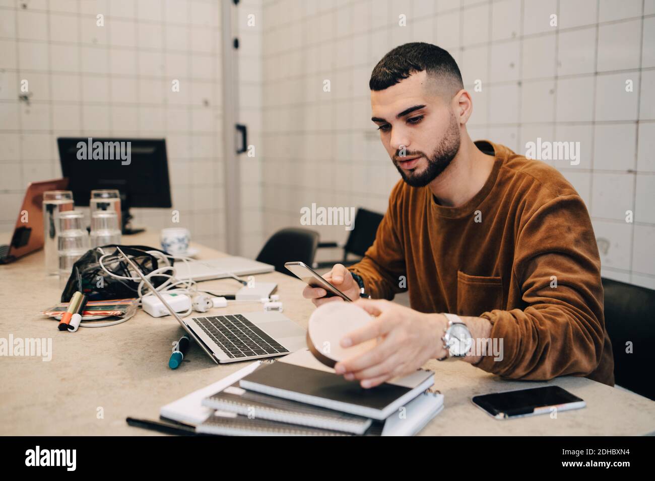 Junge männliche Hacker mit Smartphone, während die Ausrüstung zu halten Schreibtisch in kleinem Kreativbüro Stockfoto