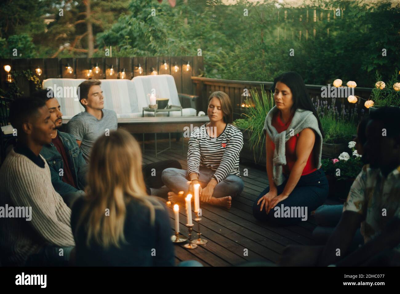 Männliche und weibliche Freunde sitzen mit geschlossenen Augen um beleuchtet Kerzen auf dem Balkon während der Gruppentherapie Stockfoto