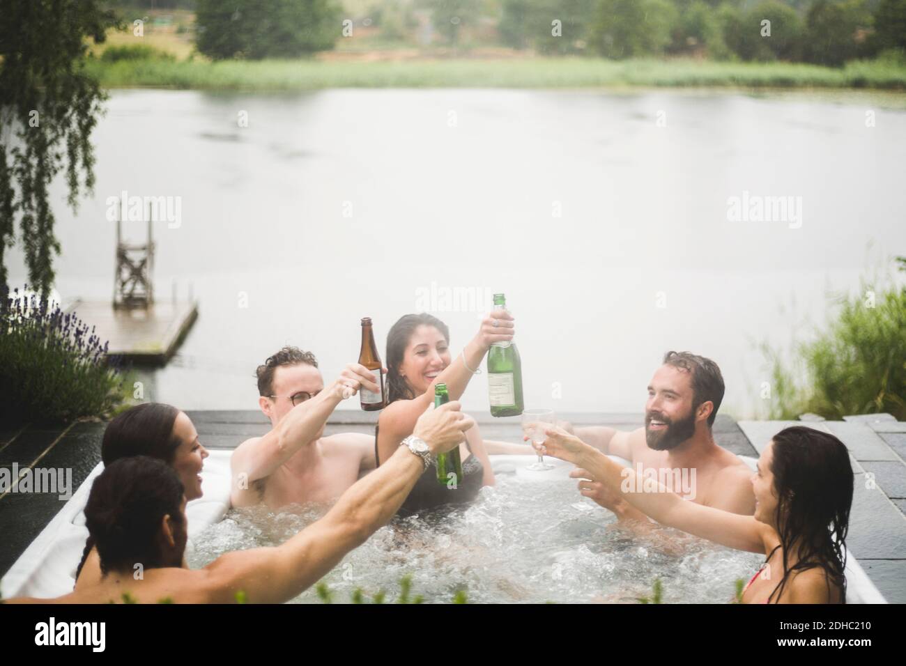 Unbeschwerte männliche und weibliche Freunde genießen Getränke im Whirlpool Gegen den See während des Wochenend-Kurzurlaubs Stockfoto