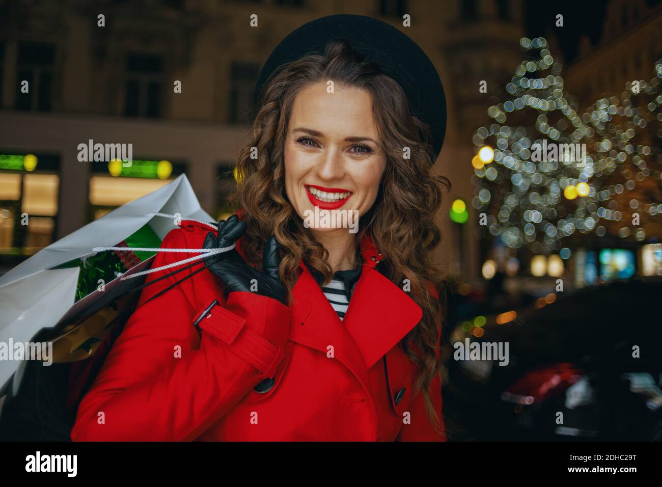 Winterspaß. Porträt von lächelnden modernen mittleren Alters Frau Shopper in roten Mantel und schwarze Baskenmütze mit Einkaufstaschen im Freien in der Stadt in der Nacht. Stockfoto