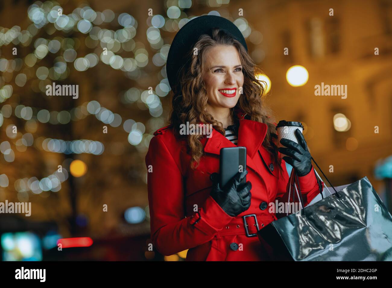 Lächelnde junge Frau Shopper in rotem Mantel und schwarzem Baskenmütze mit Einkaufstaschen, Tasse Cappuccino und Smartphone in die Ferne schauen und senden tex Stockfoto