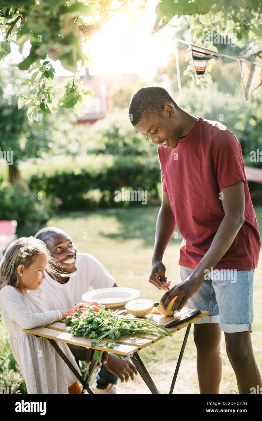 Junge schneiden Melone auf dem Tisch, während Vater und Schwester suchen Bei ihm während der Gartenparty Stockfoto