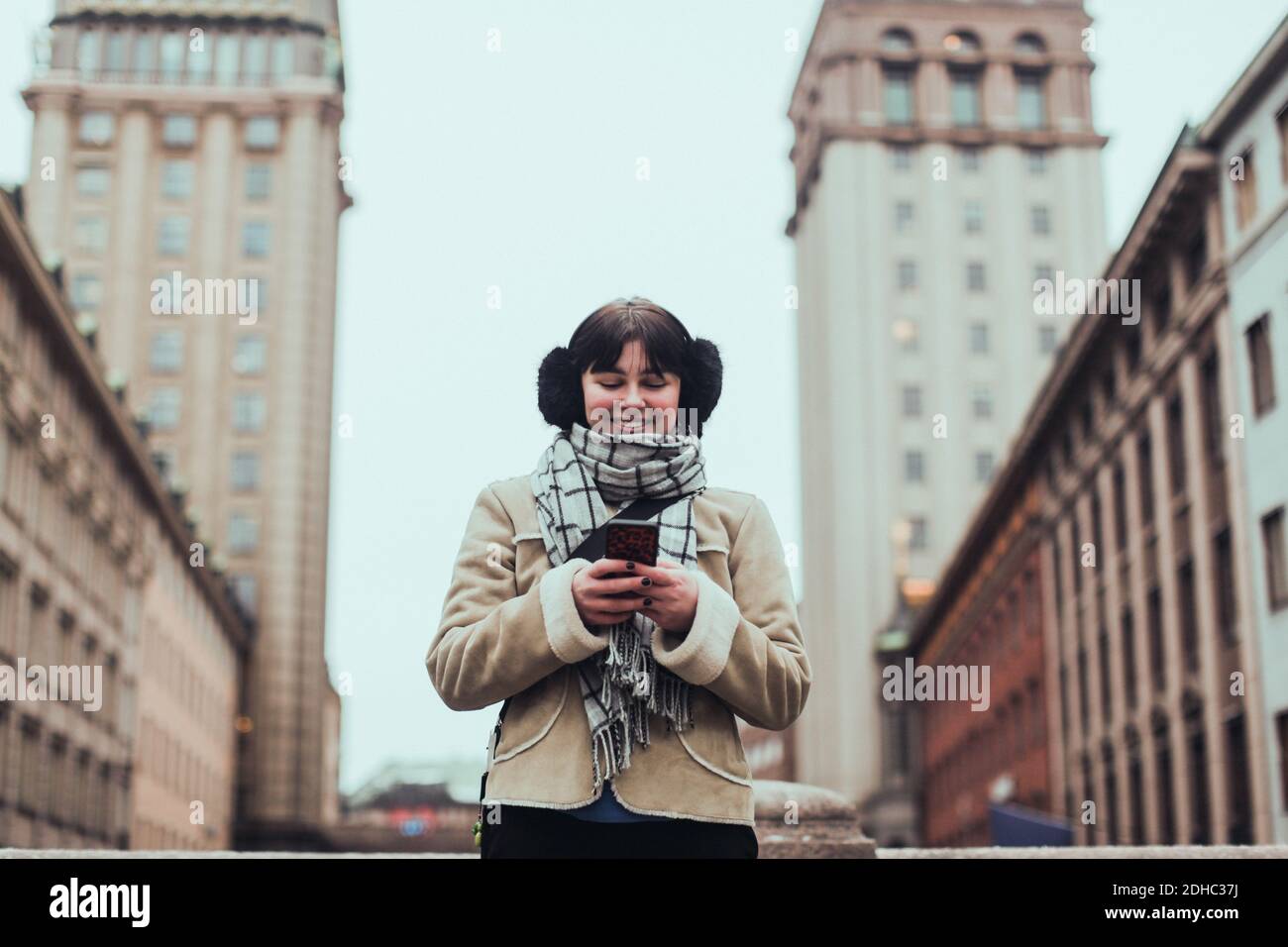 Lächelnde junge Frau in warmer Kleidung, während sie das Handy benutzt Gegen Gebäude in der Stadt Stockfoto