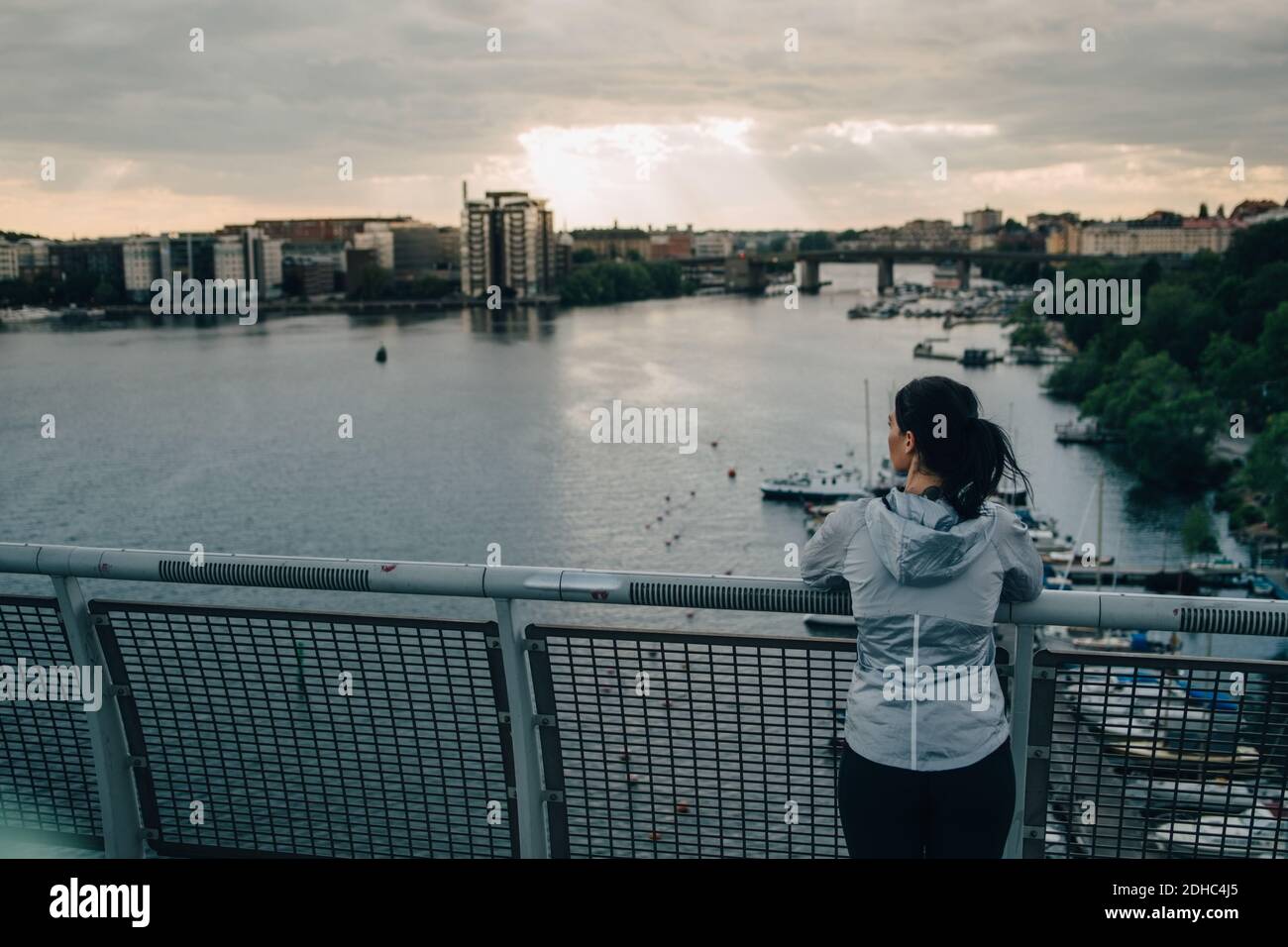 Rückansicht einer Sportlerin, die im Stehen die Stadt anschaut Auf einer Fußgängerbrücke über das Meer bei Sonnenuntergang Stockfoto