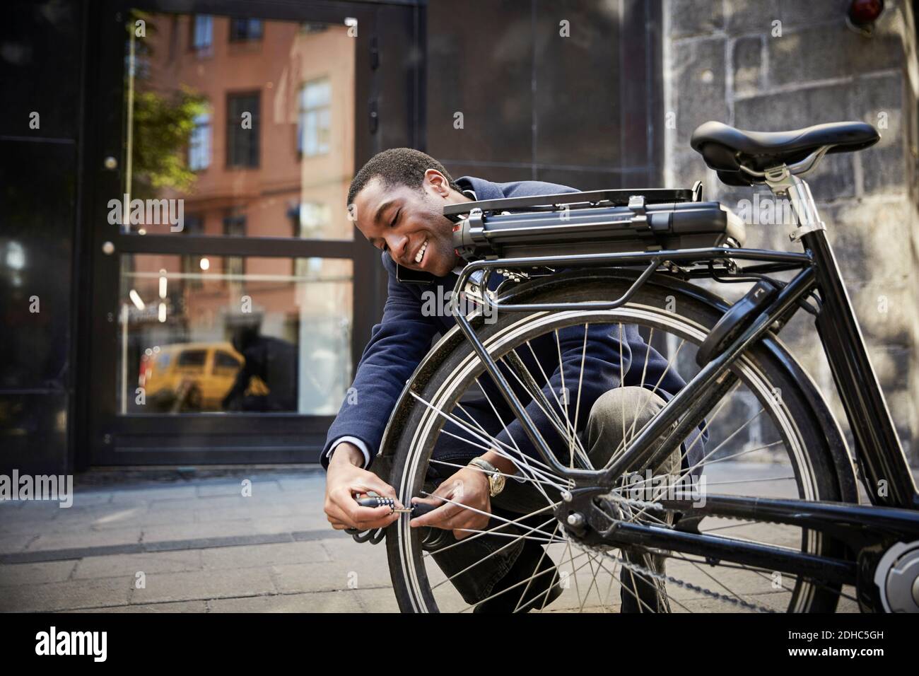 Lächelnder junger männlicher Pendler, der ein elektrisches Fahrrad verriegelt, während er sich dagegen hockend Gebäude in der Stadt Stockfoto