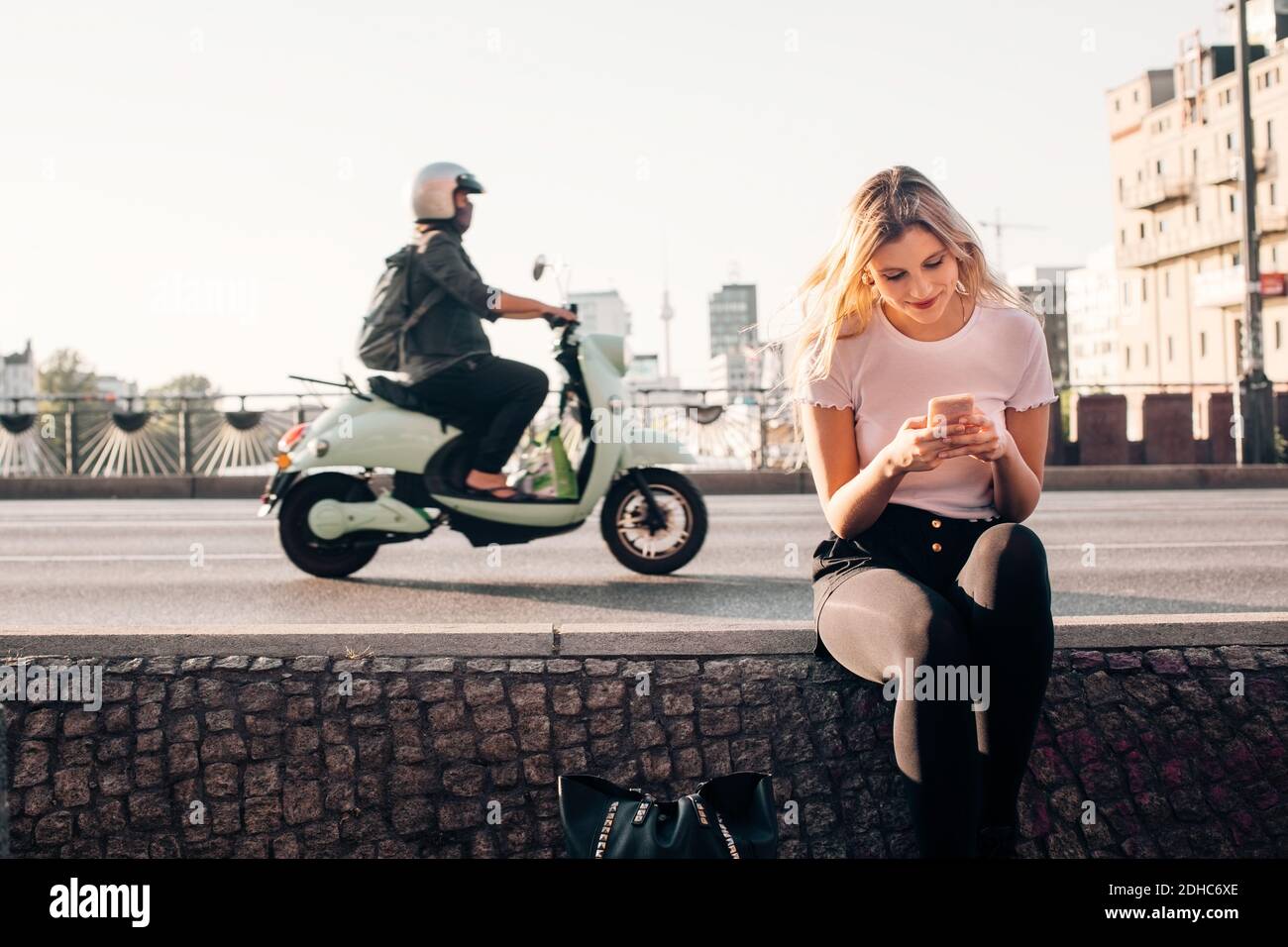 Lächelnde junge Frau mit Smartphone, während sitzen auf halten Mauer an der Straße in der Stadt Stockfoto