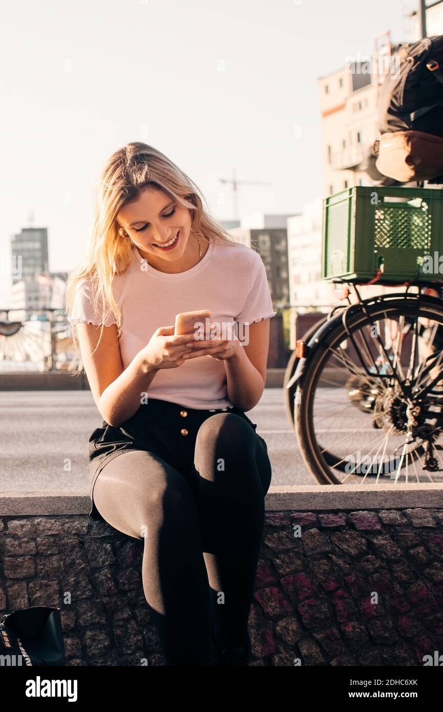Lächelnde junge Frau mit Handy, während sitzen auf halten Mauer an der Straße in der Stadt Stockfoto