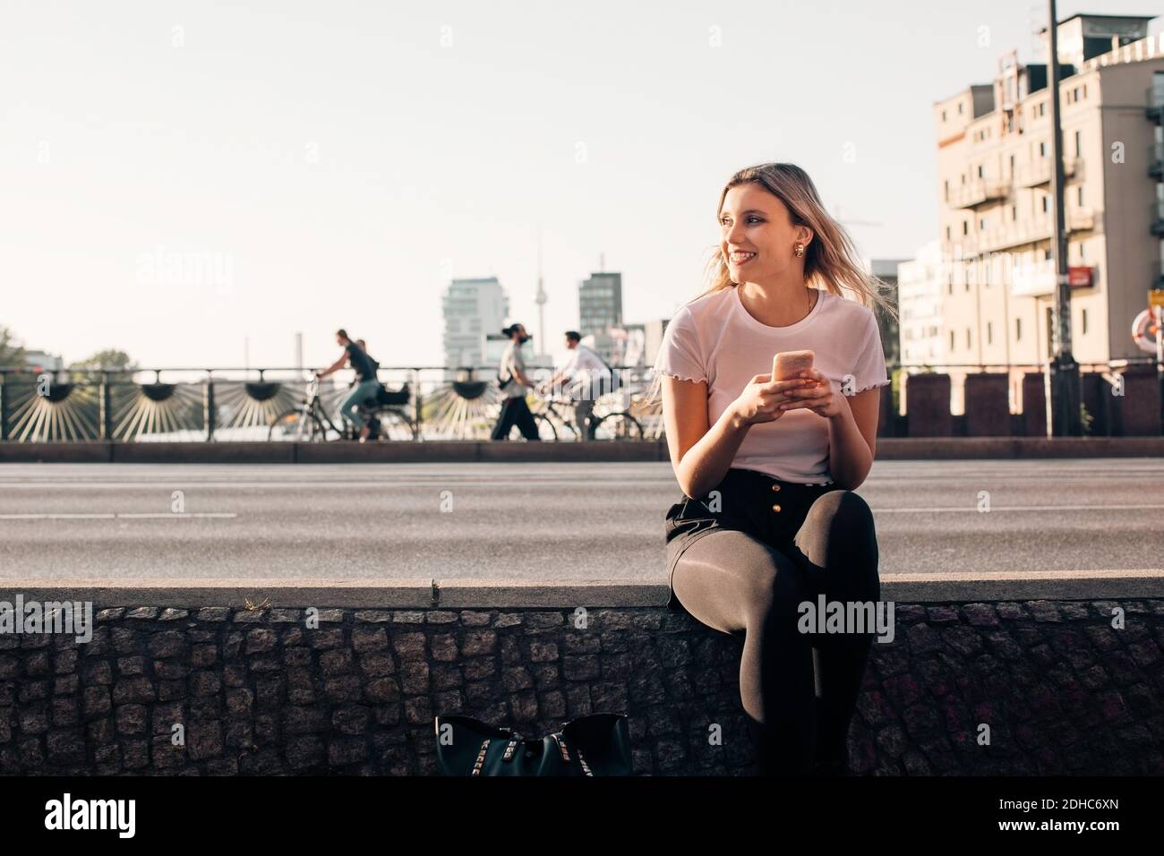 Lächelnde junge Frau hält Mobiltelefon, während sie auf Halten sitzt Mauer an der Straße Stockfoto
