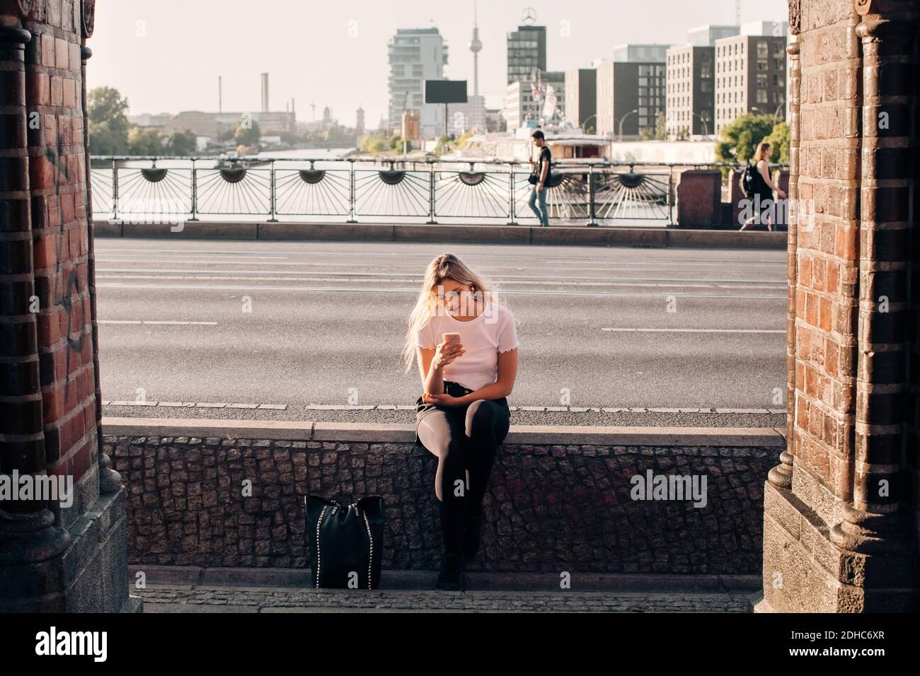 Junge Frau mit Handy, während sie auf Stützwand sitzen Auf der Straße Stockfoto