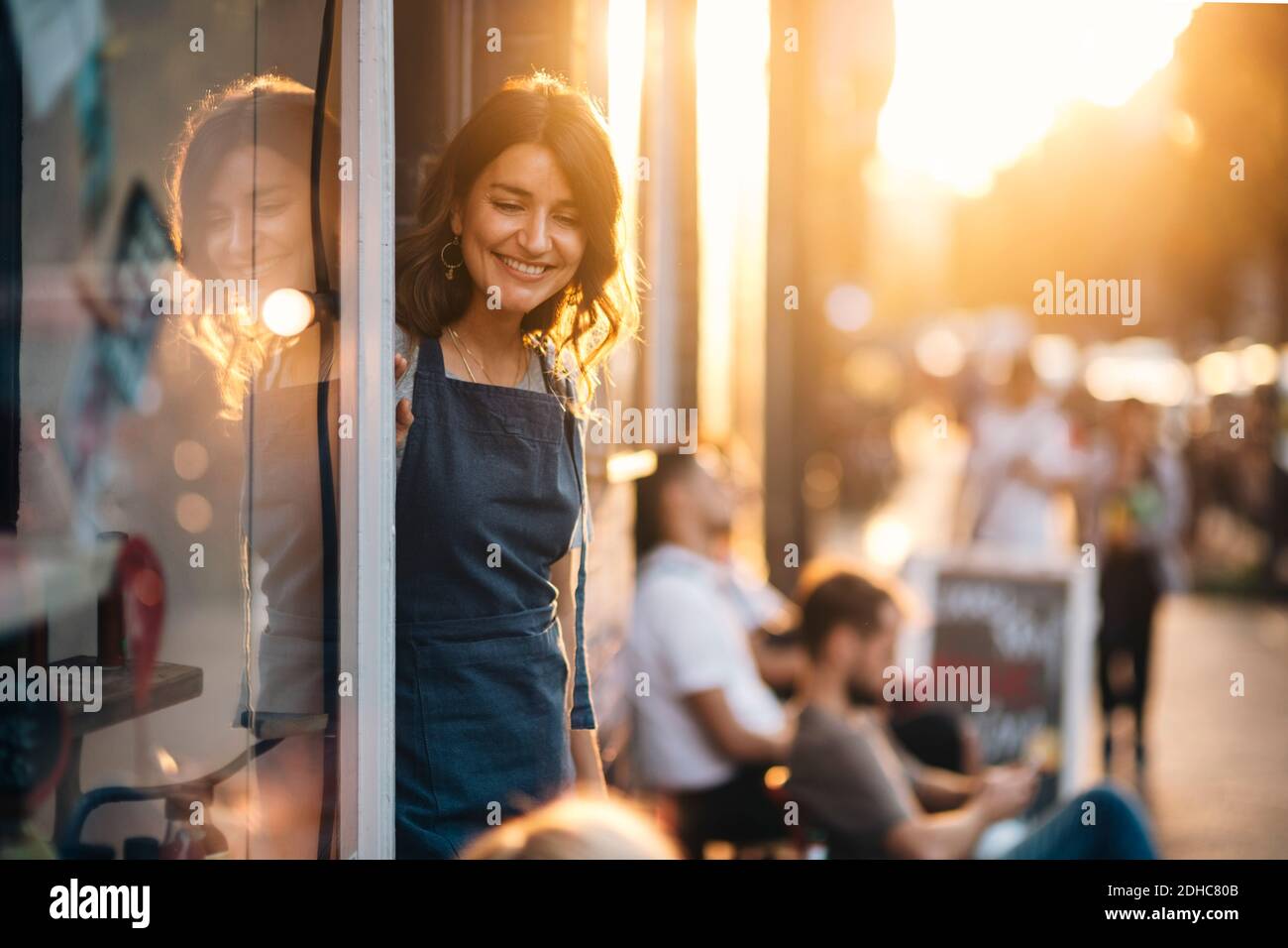 Lächelnde weibliche Besitzerin, die unten schaut, während sie am Eingang von steht Feinkostgeschäft Stockfoto