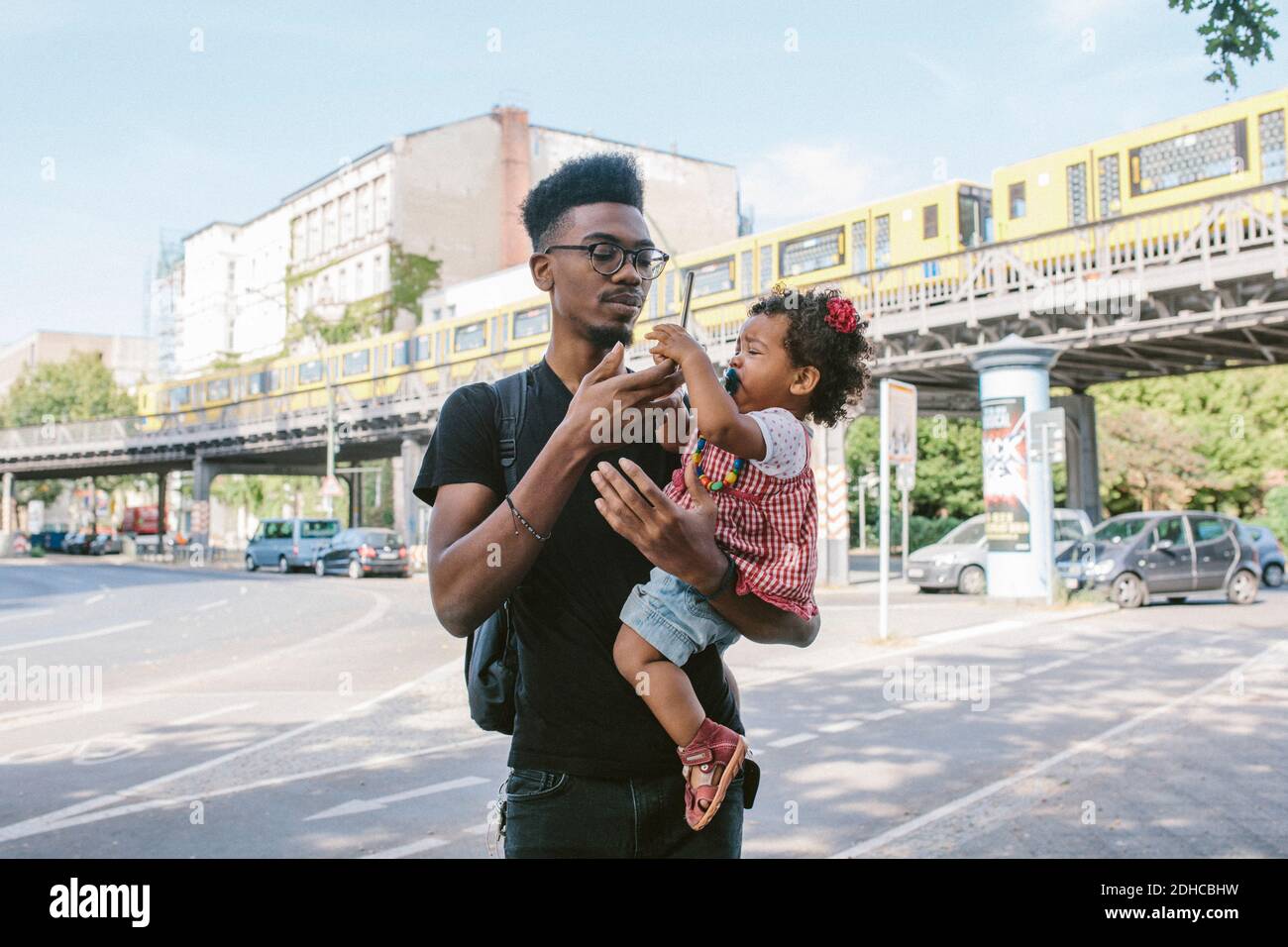 Junger Mann, der seine Tochter mit dem Mobiltelefon trägt, während er auf ihm steht Straße gegen Eisenbahnbrücke Stockfoto