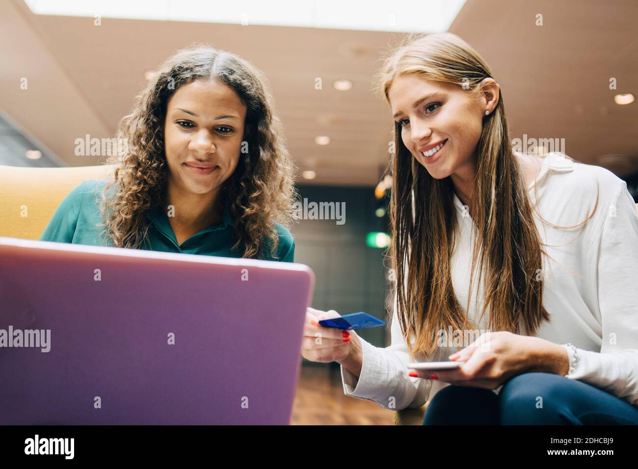 Flache Ansicht von lächelnden weiblichen Kollegen aus verschiedenen Ethnien, die einen Laptop benutzen Beim Sitzen in der Flughafenlobby Stockfoto