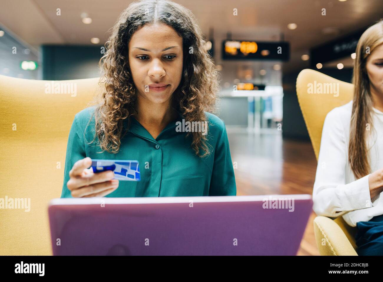 Selbstbewusste Geschäftsfrau mit Kreditkarte, während sie den Laptop von einem Kollegen benutzt In der Flughafenlobby Stockfoto