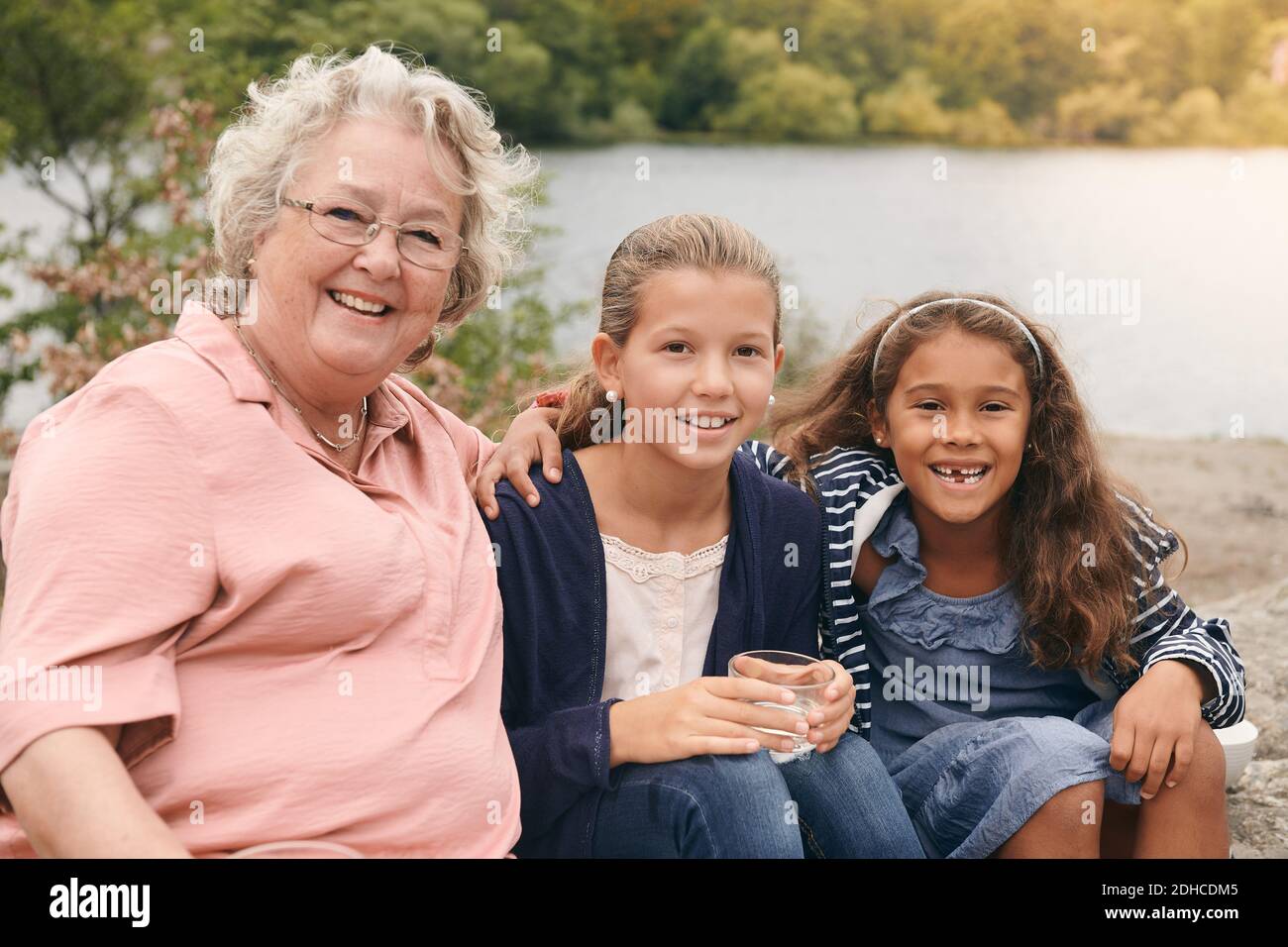 Porträt von lächelnden Enkelinnen und Großmutter sitzen am Seeufer in parken Stockfoto