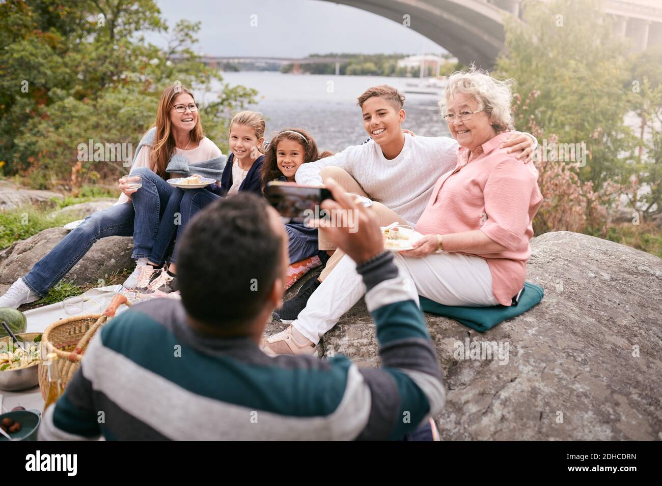 Mann fotografiert glückliche Familie mit Essen auf Felsen im Park Während des Picknicks Stockfoto