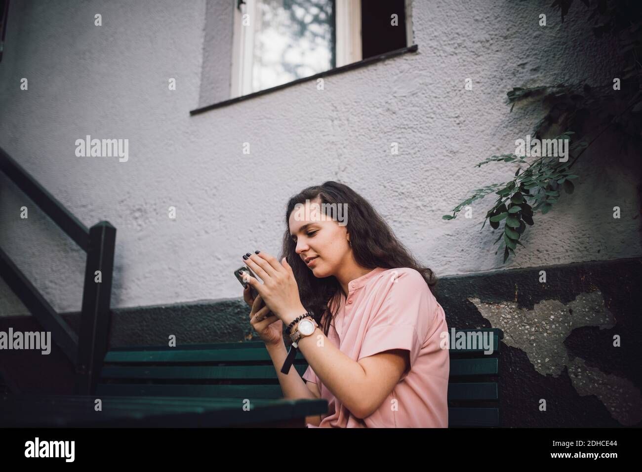 Junge Frau mit Handy, während sie auf der Bank draußen sitzen Haus Stockfoto