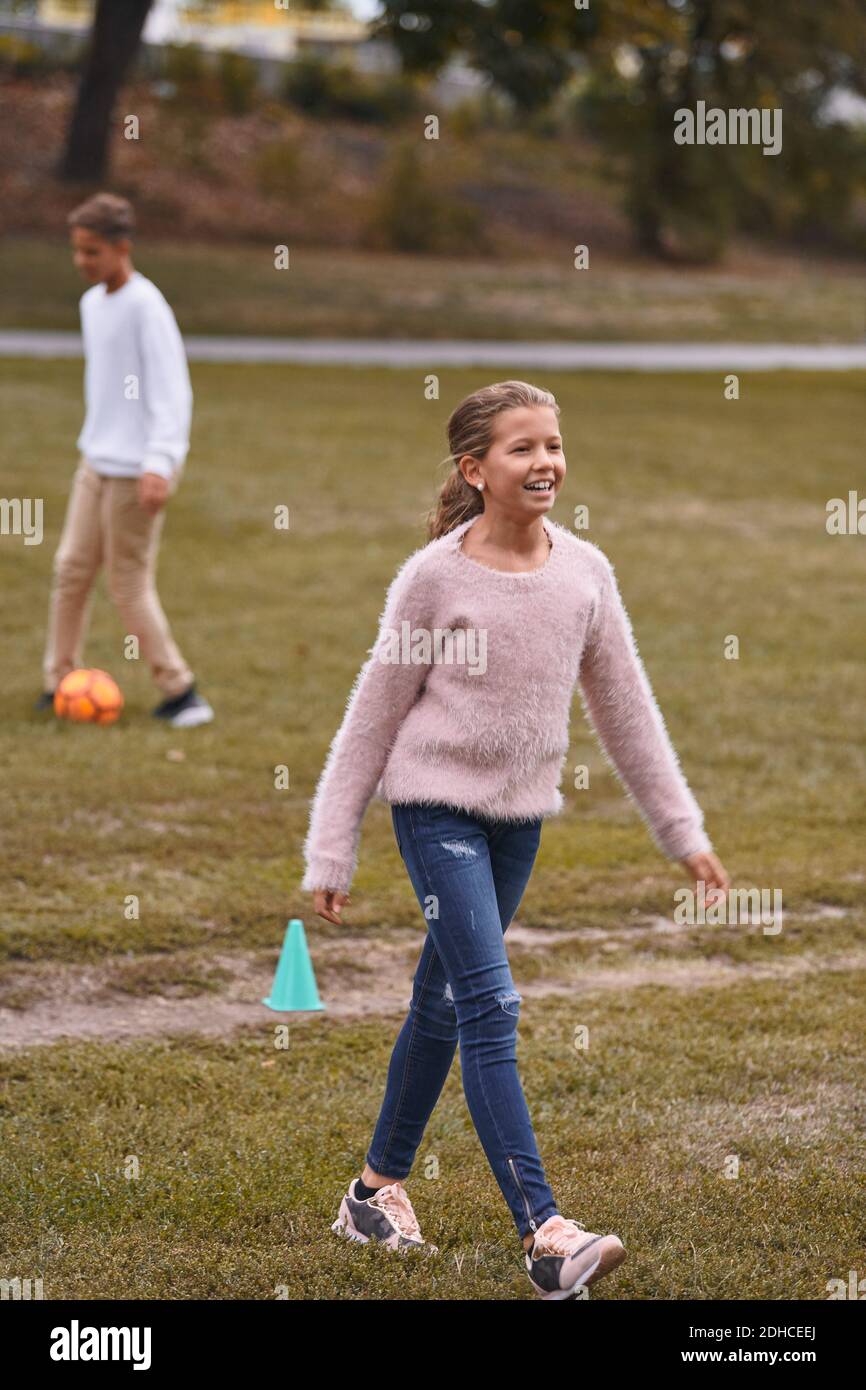 Lächelnde Schwester, die auf dem Grasfeld läuft, während Bruder Fußball spielt Im Hintergrund im Park Stockfoto