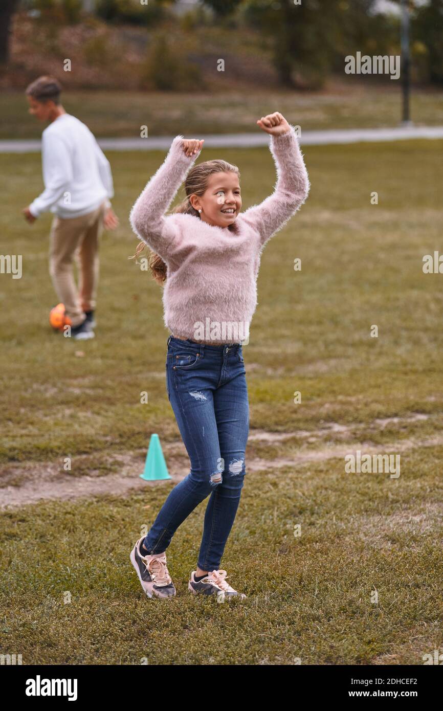 Lächelnde Schwester mit erhobenen Armen stehen auf grasbewachsenen Feld, während Bruder spielt Fußball im Hintergrund im Park Stockfoto