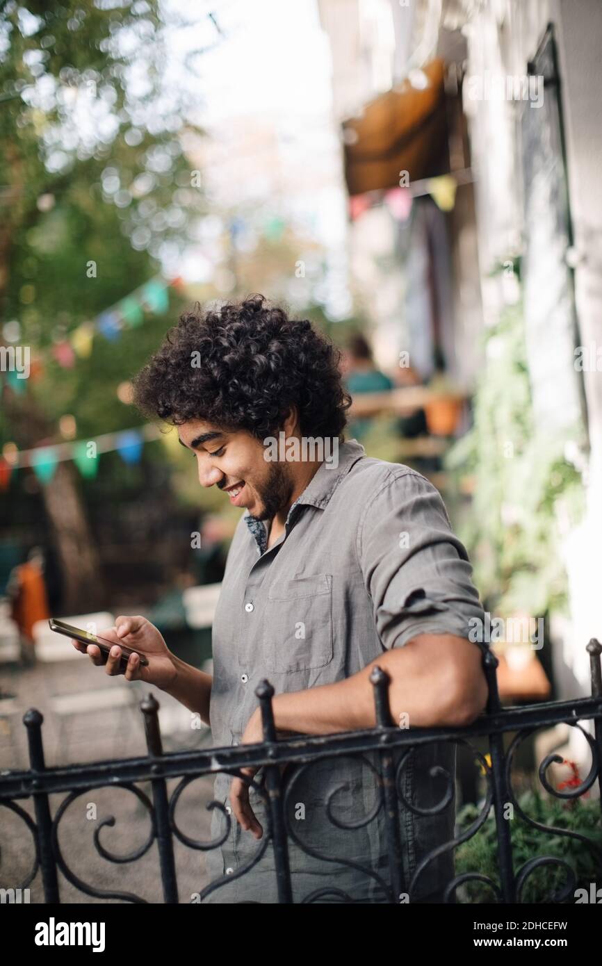 Lächelnder junger Mann mit Handy, während er sich auf Geländer stützt Im Hinterhof Stockfoto