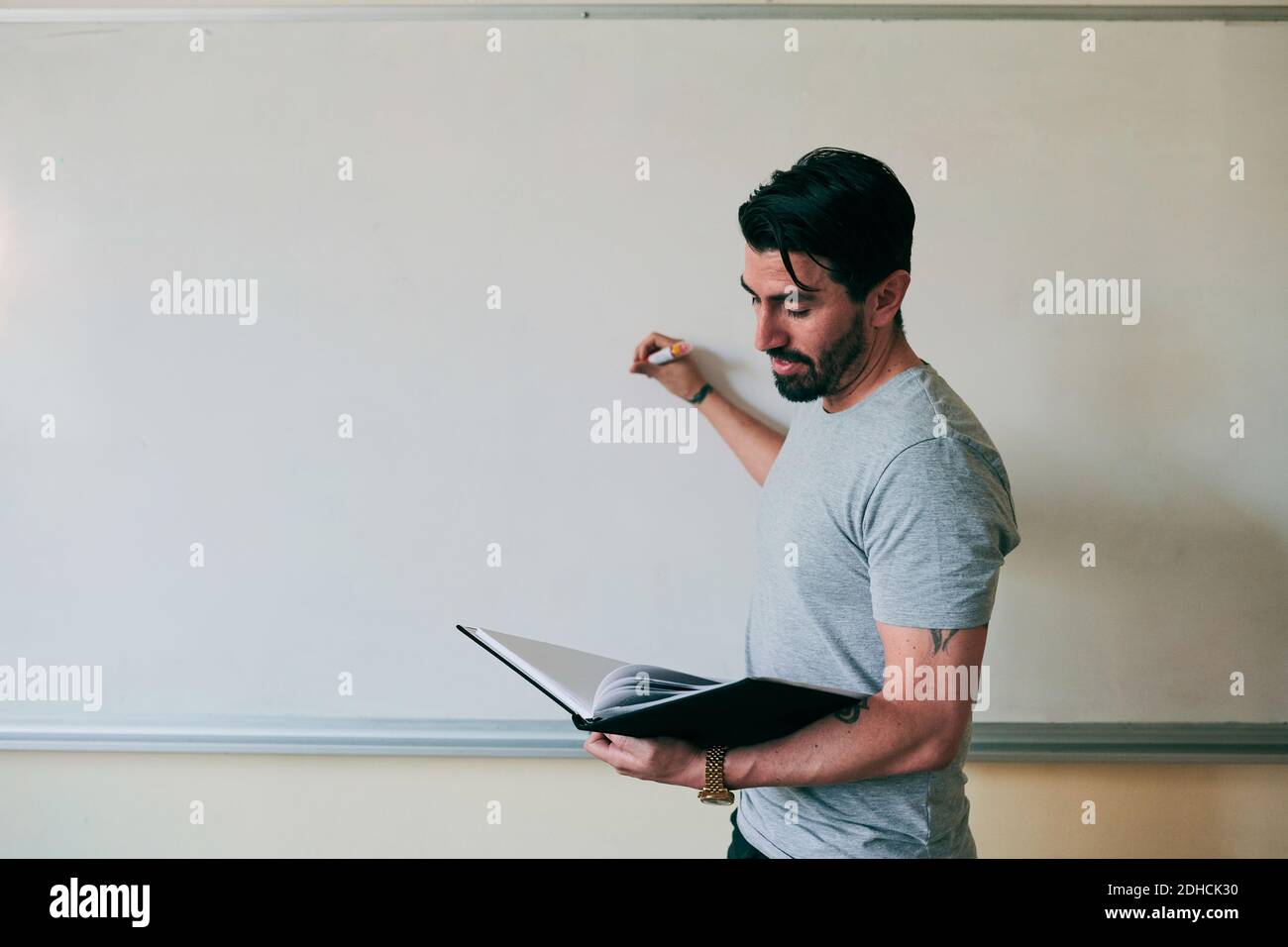 Selbstbewusster männlicher Lehrer hält Buch beim Schreiben auf Whiteboard Stockfoto