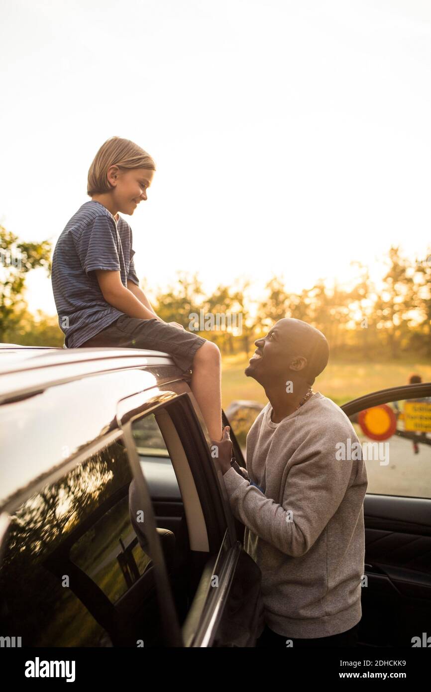 Lächelnder Mann, der während der Fahrt Mädchen auf dem Autodach ansieht Sonnenuntergang im Park Stockfoto