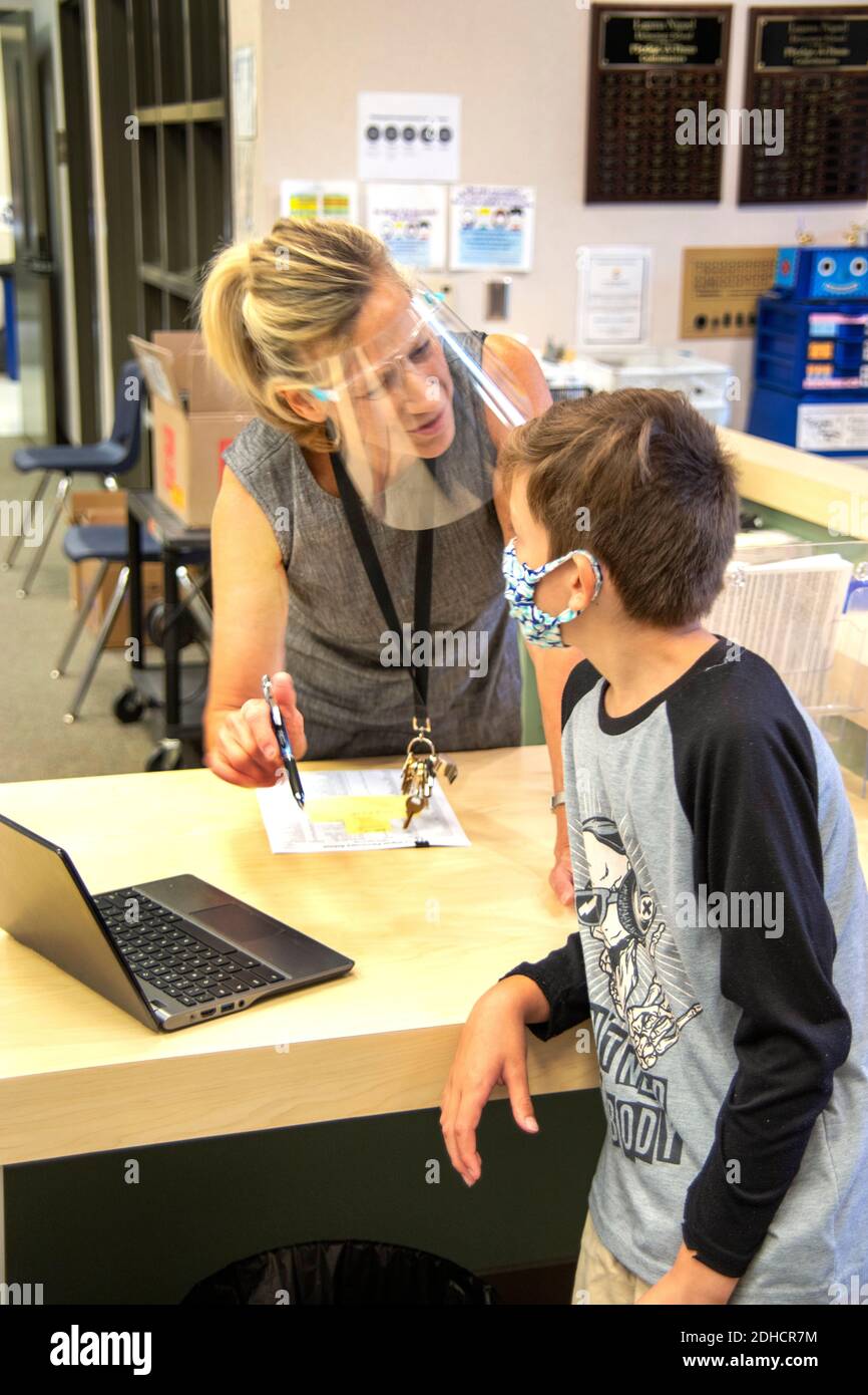 Der Rezeptionist einer kalifornischen Grundschule, der aufgrund der Coronavirus-Pandemie maskiert wurde, hilft einem Schüler, der Schwierigkeiten mit seinem Acer hat Stockfoto