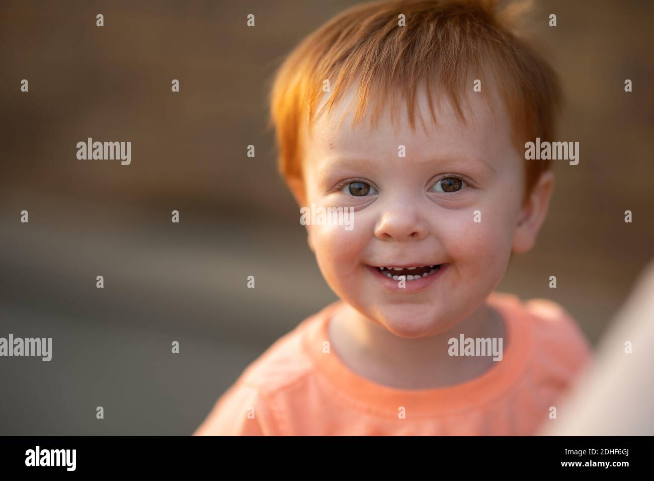 Ein entzückender, rothaariger kleiner Junge in einem orangefarbenen Hemd (mein Sohn) lächelt bei einem warmen Licht am späten Nachmittag an der Kamera. Stockfoto